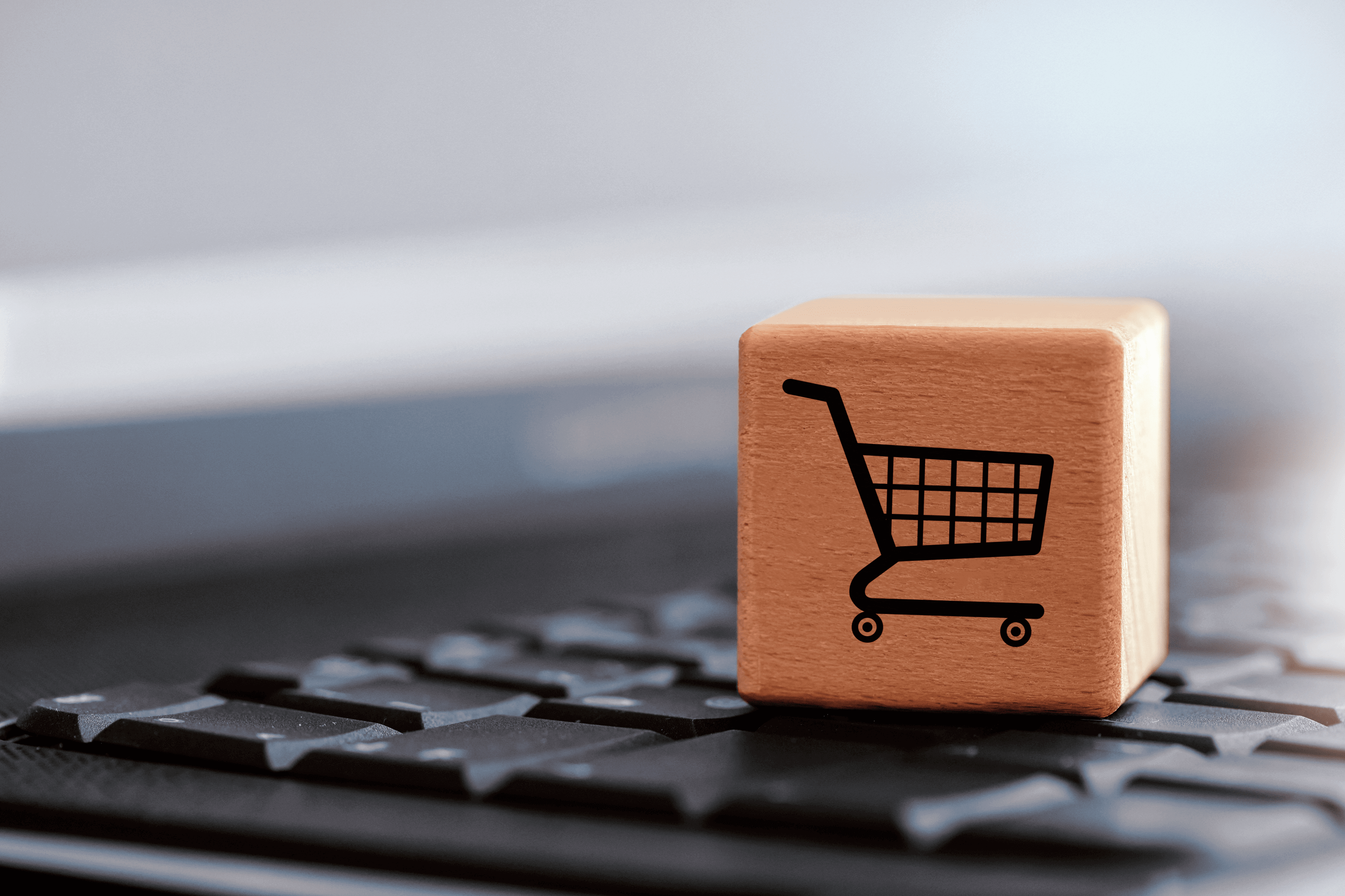 Wooden cube with a black shopping cart icon placed on a computer keyboard, symbolizing e-commerce or online shopping.